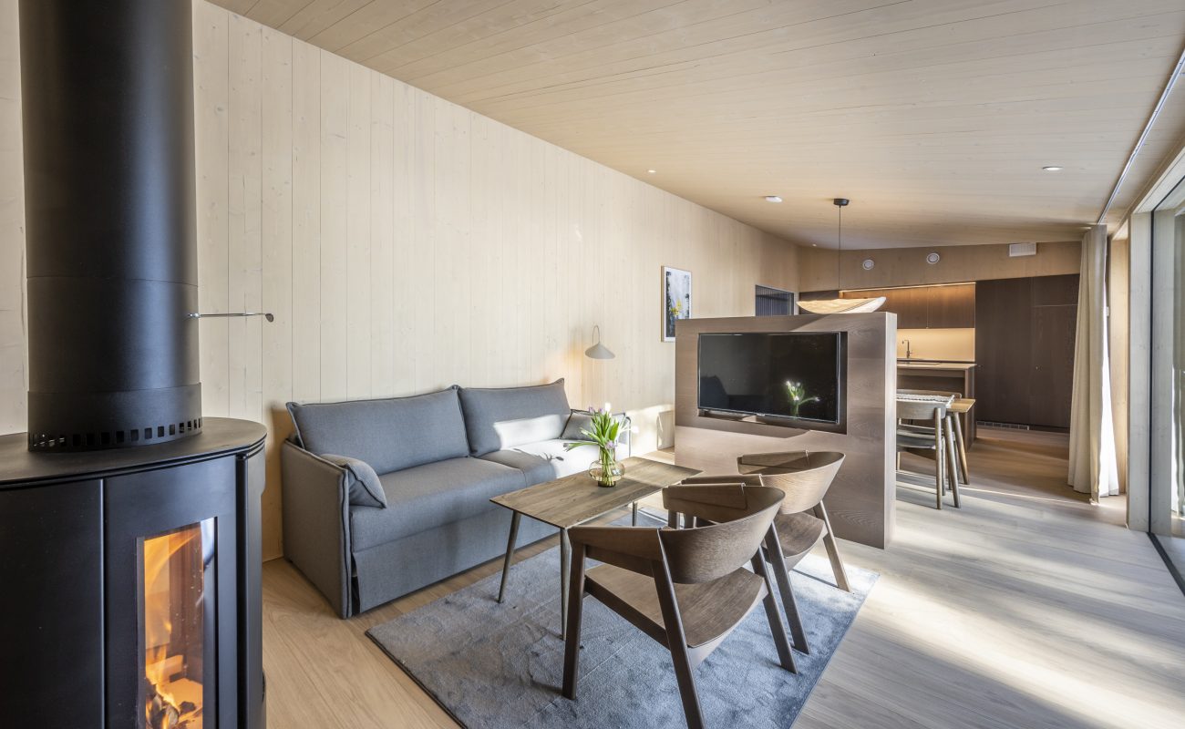 A cozy living room featuring a fireplace, sofa, wooden table, and chairs, leads to a modern kitchen.