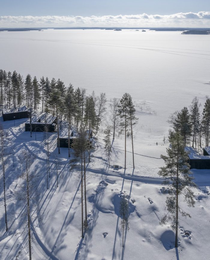 Talvisessa maisemassa on luminen järvi ja metsän laidassa useita mustia rakennuksia.