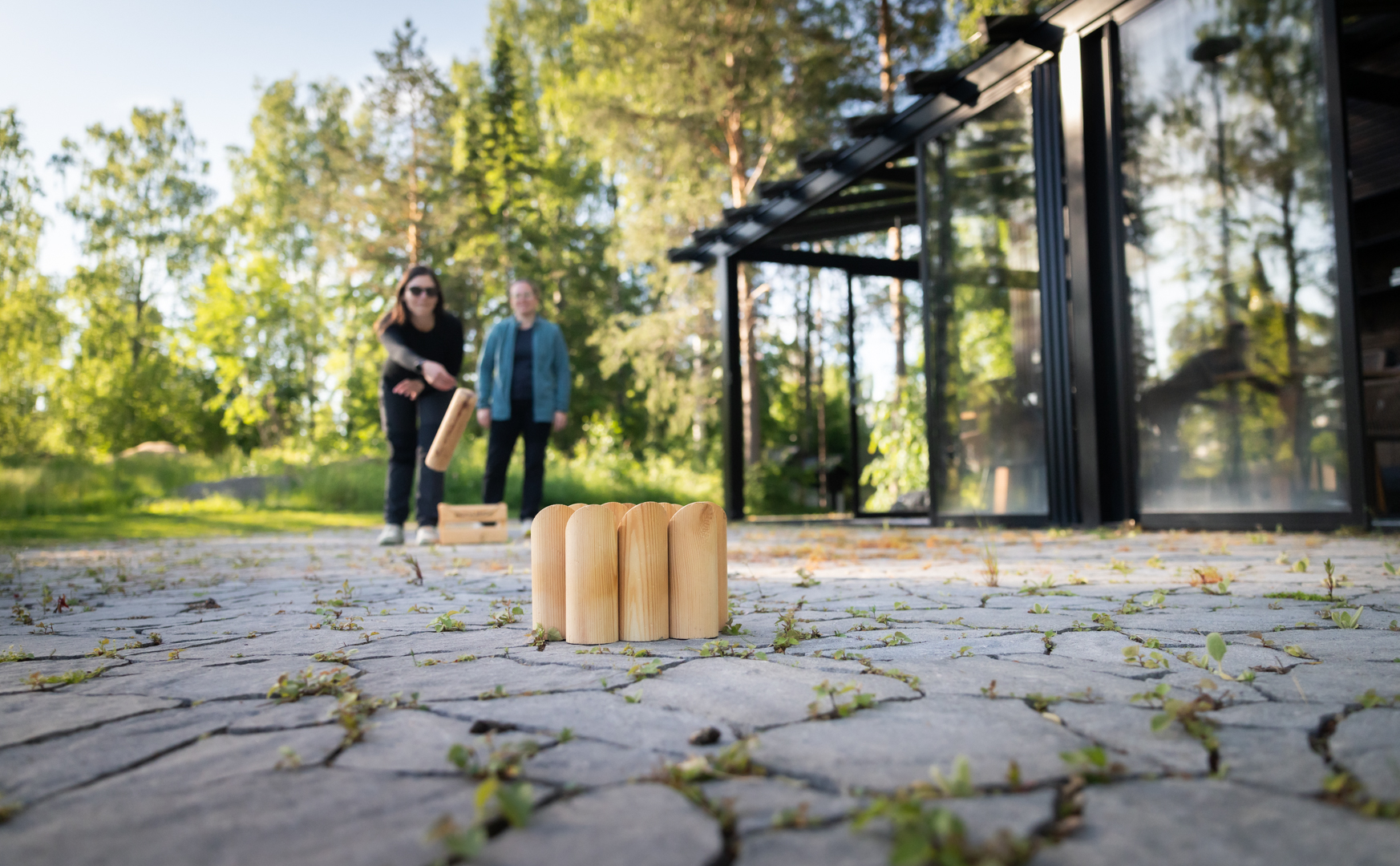 Kaksi henkilöä pelaa ulkona mölkkyä vihreän luonnon keskellä modernin rakennuksen edustalla.