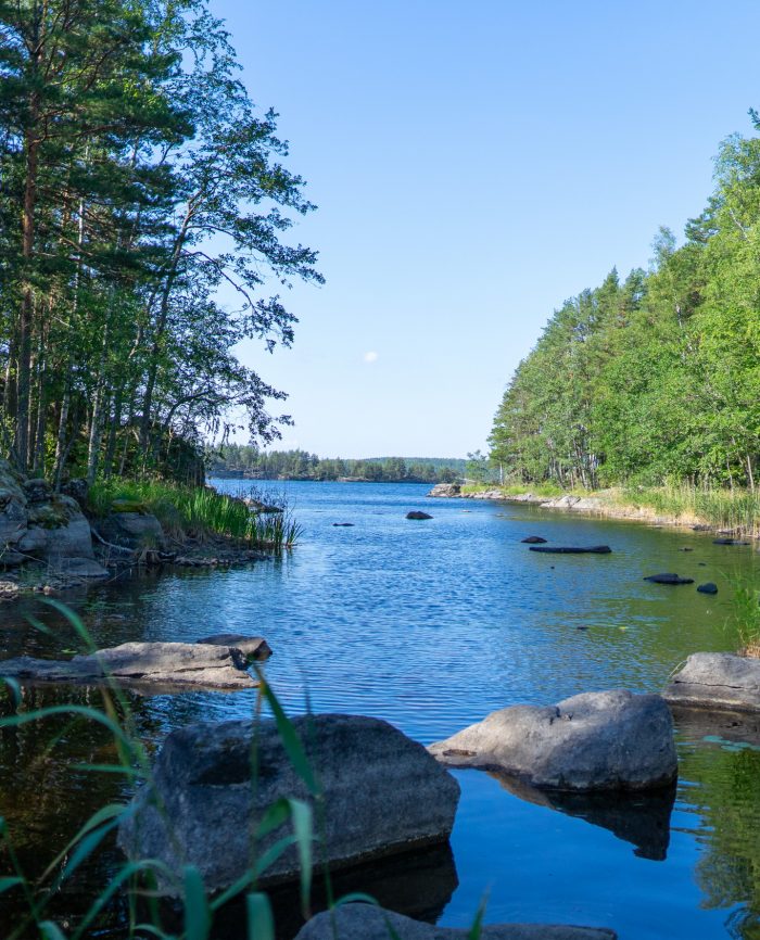 Tyyni järvimaisema, jossa on kiviä, vihreitä puita ja sininen taivas.