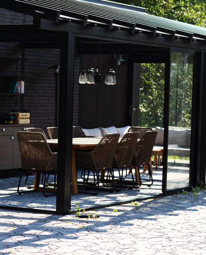 Tyylikäs, moderni lasiterassi, jossa on avokeittiö, ruokapöytä ja mukavat tuolit luonnonläheisessä ympäristössä.