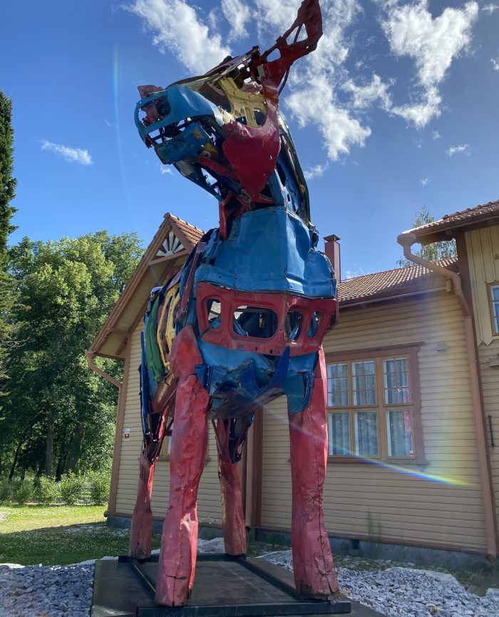 A large, colorful, and modern sculpture of a cow made from recycled car parts, located next to a wooden building.