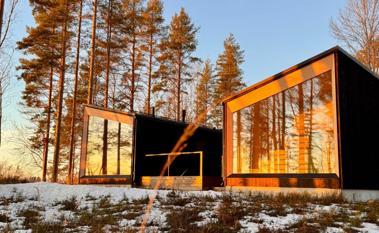 Two modern structures with large glass windows reflecting the sunset in a winter forest.