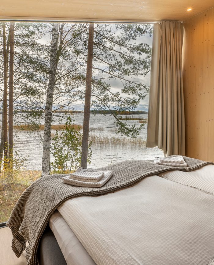 Moderni makuuhuone, jossa on lasiseinä, josta avautuu näkymä järvelle ja metsiin.