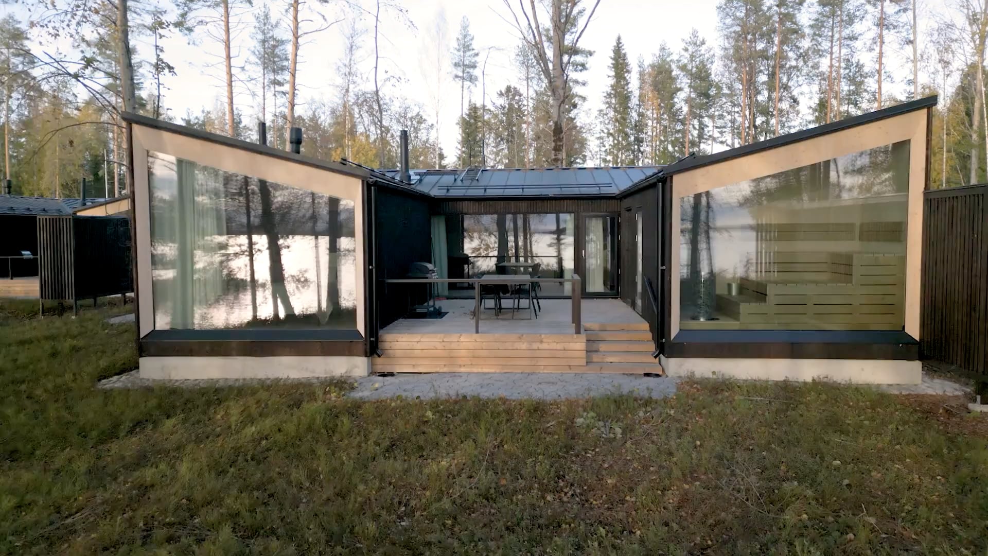 A modern cabin with large glass walls is situated in a forested area by the lakeshore.