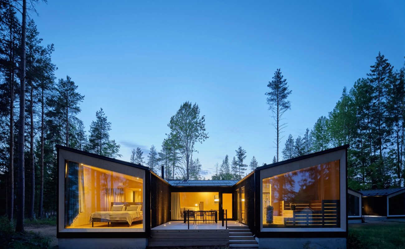 A modern cabin opens to a forested environment, illuminated under the evening sky.